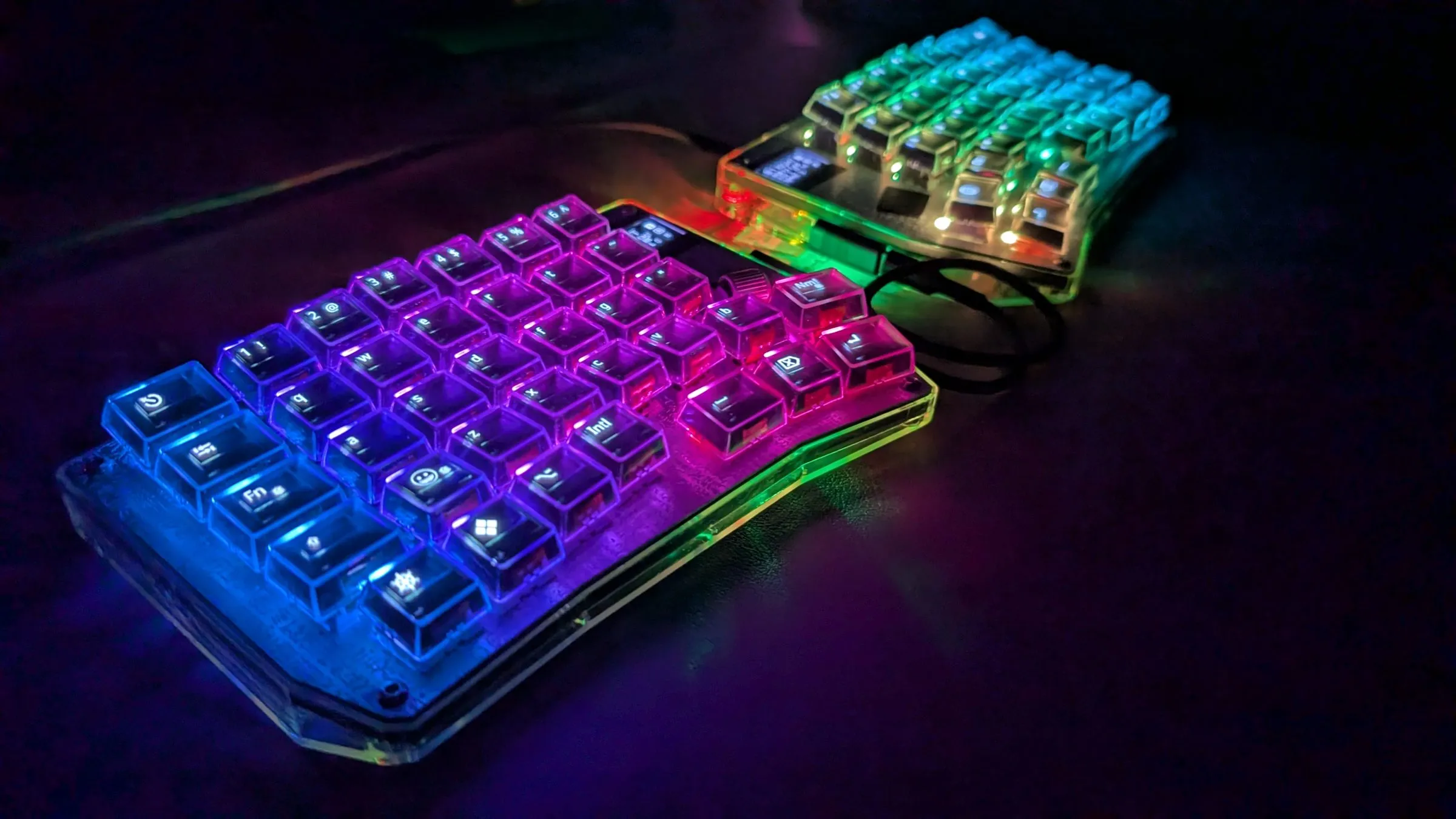 Both PolyKybd halves in the dark, each key lit by its own RGB LED in a rainbow gradient running from blue through purple to magenta, with the OLED legends readable through the transparent keycaps.